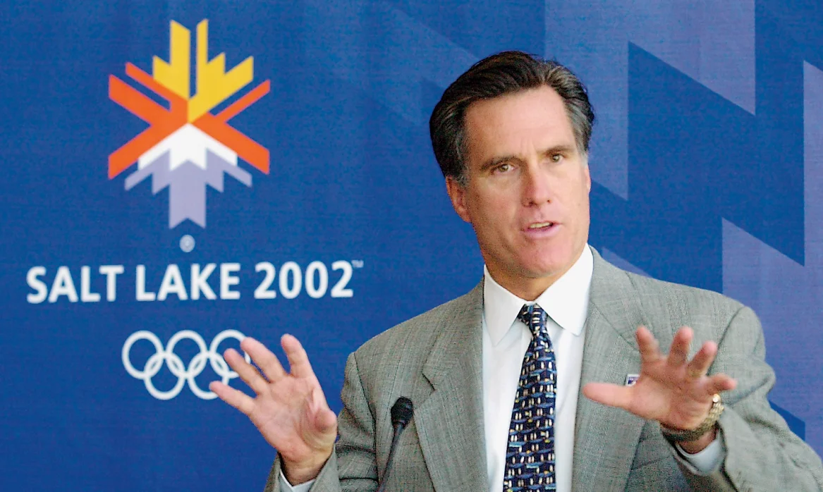 Mitt Romney speaking in front of a sign that reads "Salt Lake 2002" with the Olympic rings beneath.