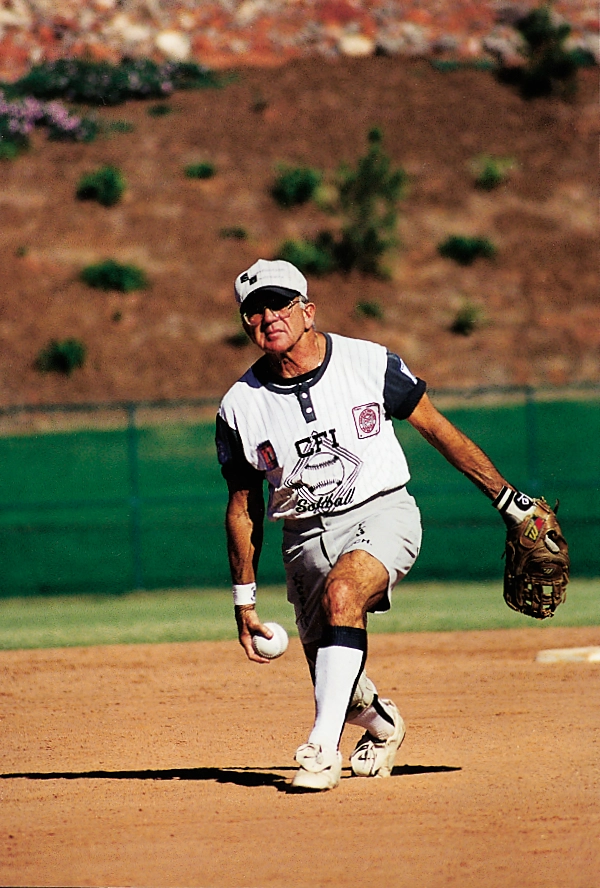 A senior athlete in a white uniform throws a softball on a dirt field.
