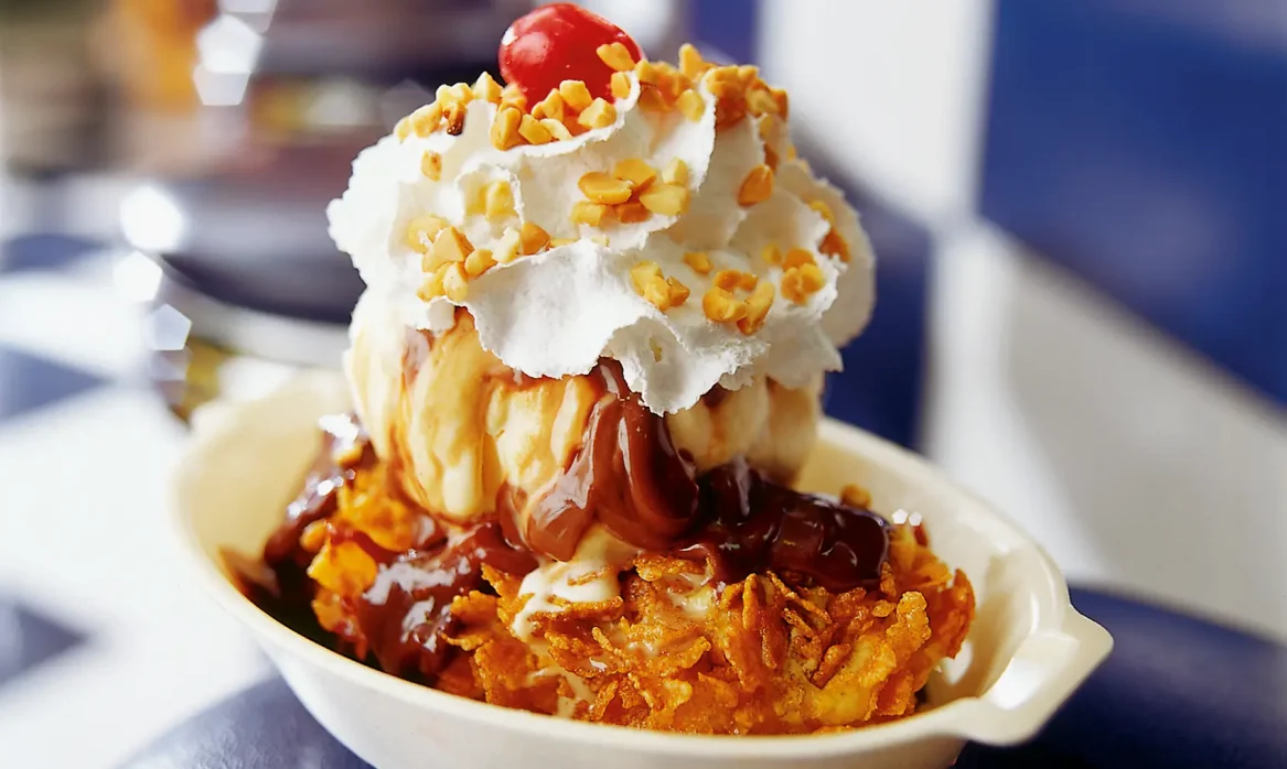 Deep-fried Milky Way bar sundae topped with scoops of ice cream, chocolate sauce, whipped cream, chopped nuts, and a cherry in a white bowl.