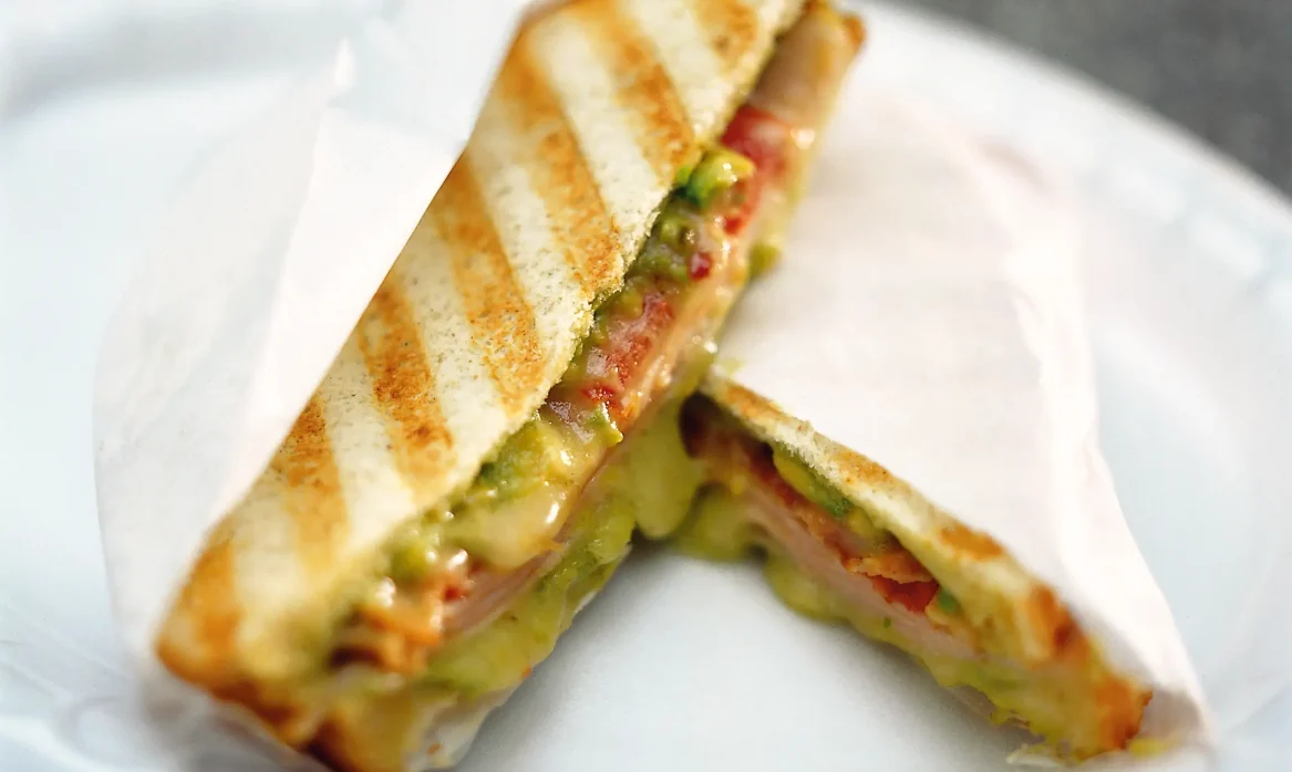 Grilled Montana panino cut into two triangular halves, with melted cheese, guacamole, tomato, turkey, and bacon visible between toasted bread.