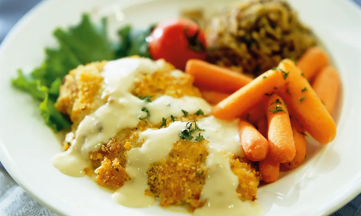 Herb-crusted chicken served with glazed baby carrots, leafy greens, a cherry tomato, and a mound of rice on a white plate, with another entrée blurred in the background.