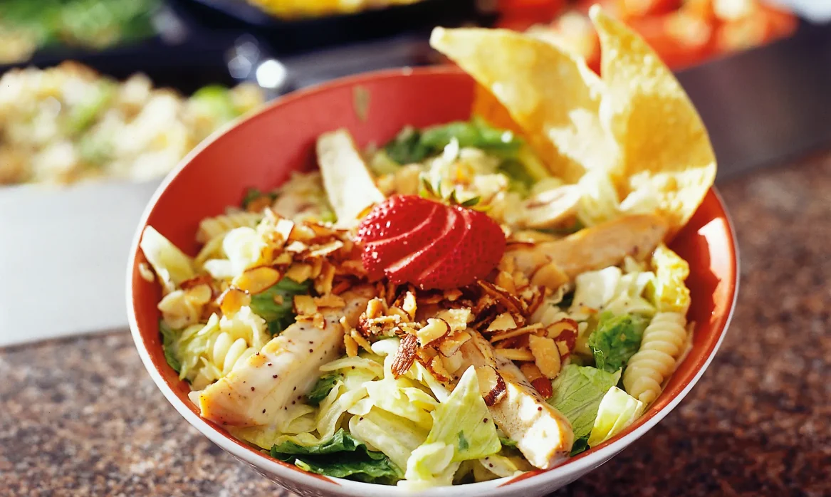 Bowl of pasta poppyseed salad with leafy greens, spiral pasta, sliced chicken, strawberries, almonds, and tortilla chips.