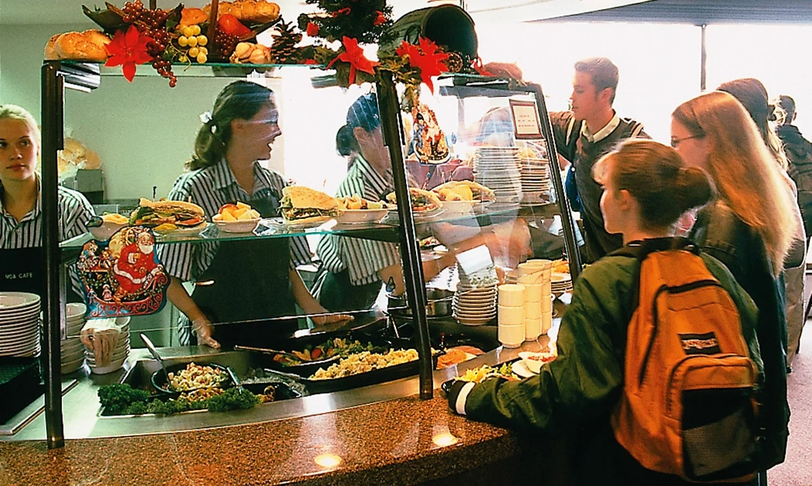 Students stand in line in front of a lunch counter where workers are serving food.