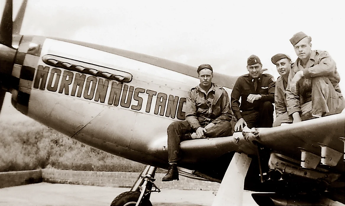 Four World War II pilots sit on the wing of a fighter plane that has the name "Mormon Mustang" painted on the side.