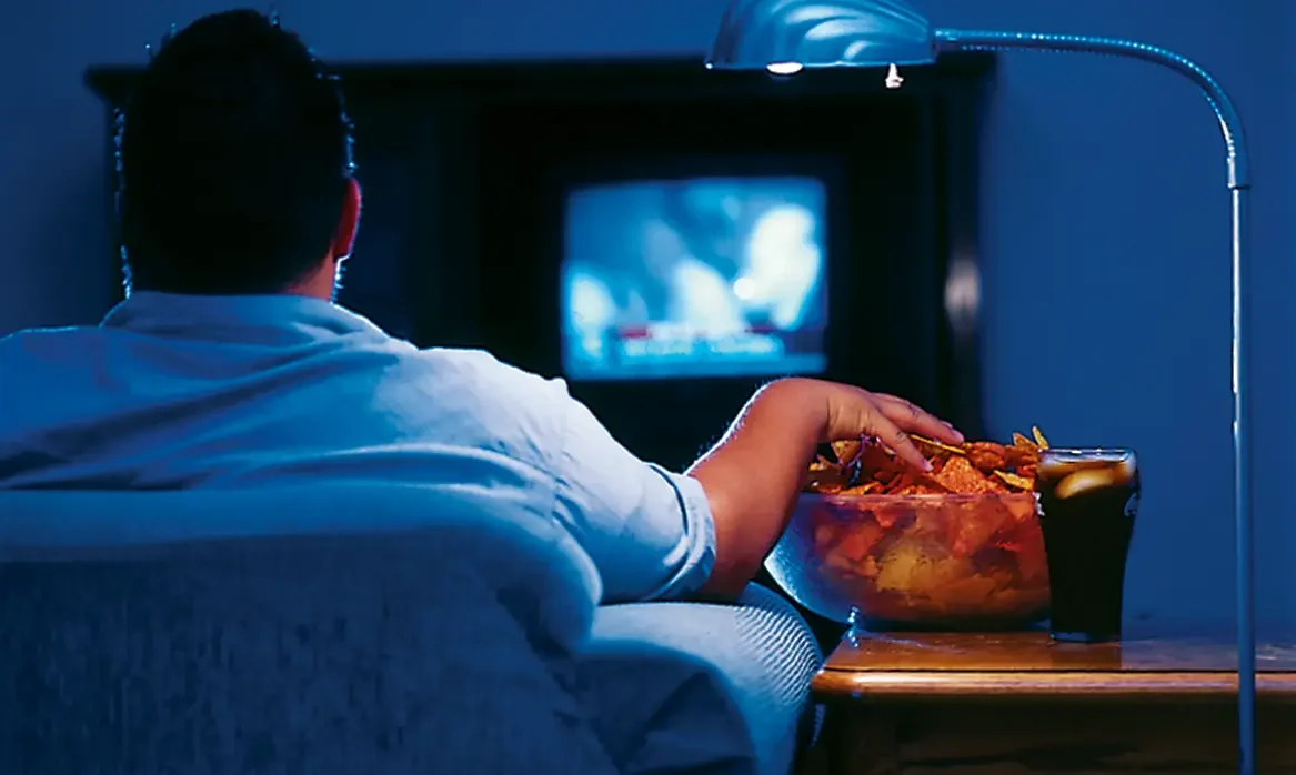 Person viewed from behind sits in a darkened room watching television while reaching into a large bag of chips on a side table.