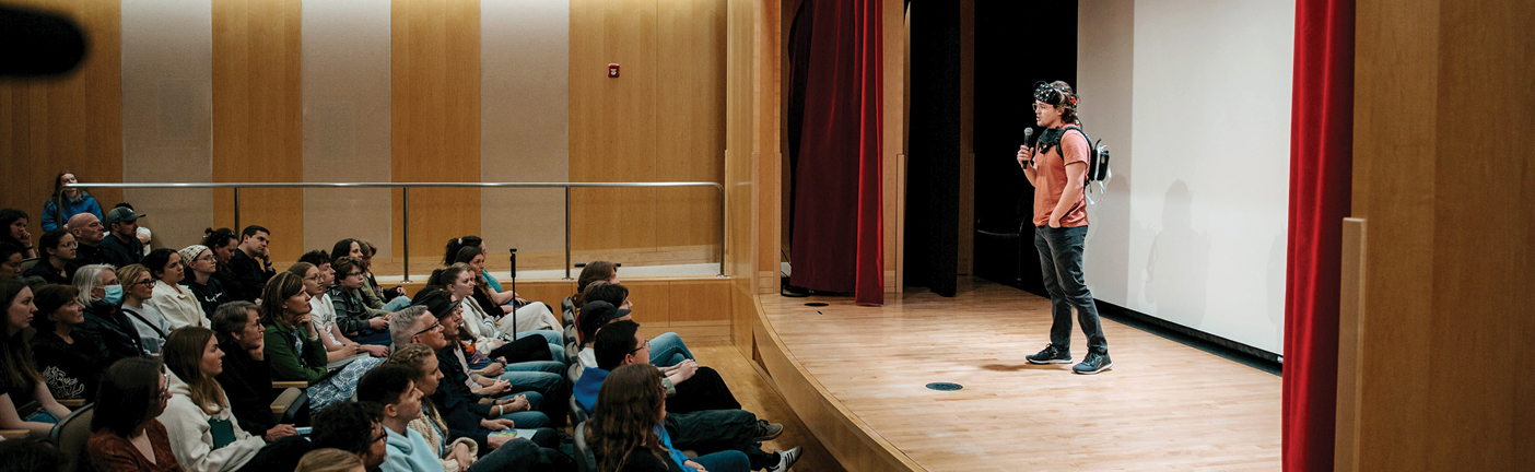 Wide view of an auditorium where an autistic storyteller speaks onstage to a seated audience during BYU’s Autistic Story SLAM; some participants wear brain-scanning devices as part of a double empathy problem study.