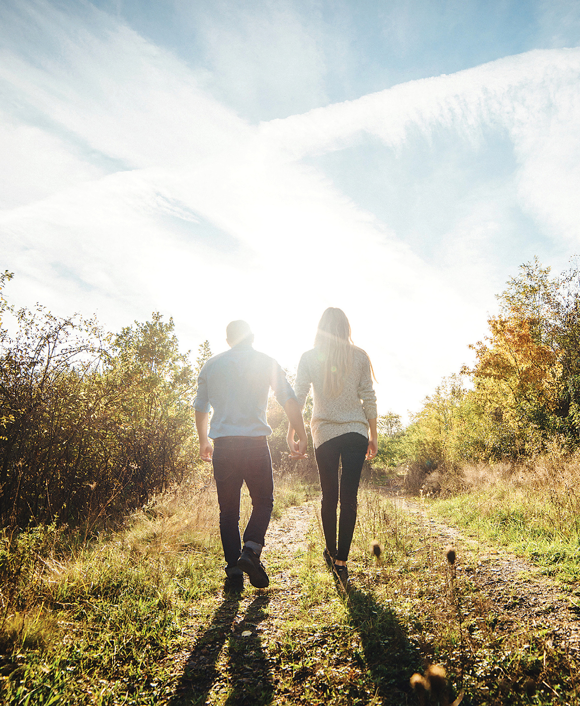 Photo of a couple walking hand in hand away from the camera along a sunlit, wooded trail surrounded by autumn foliage.