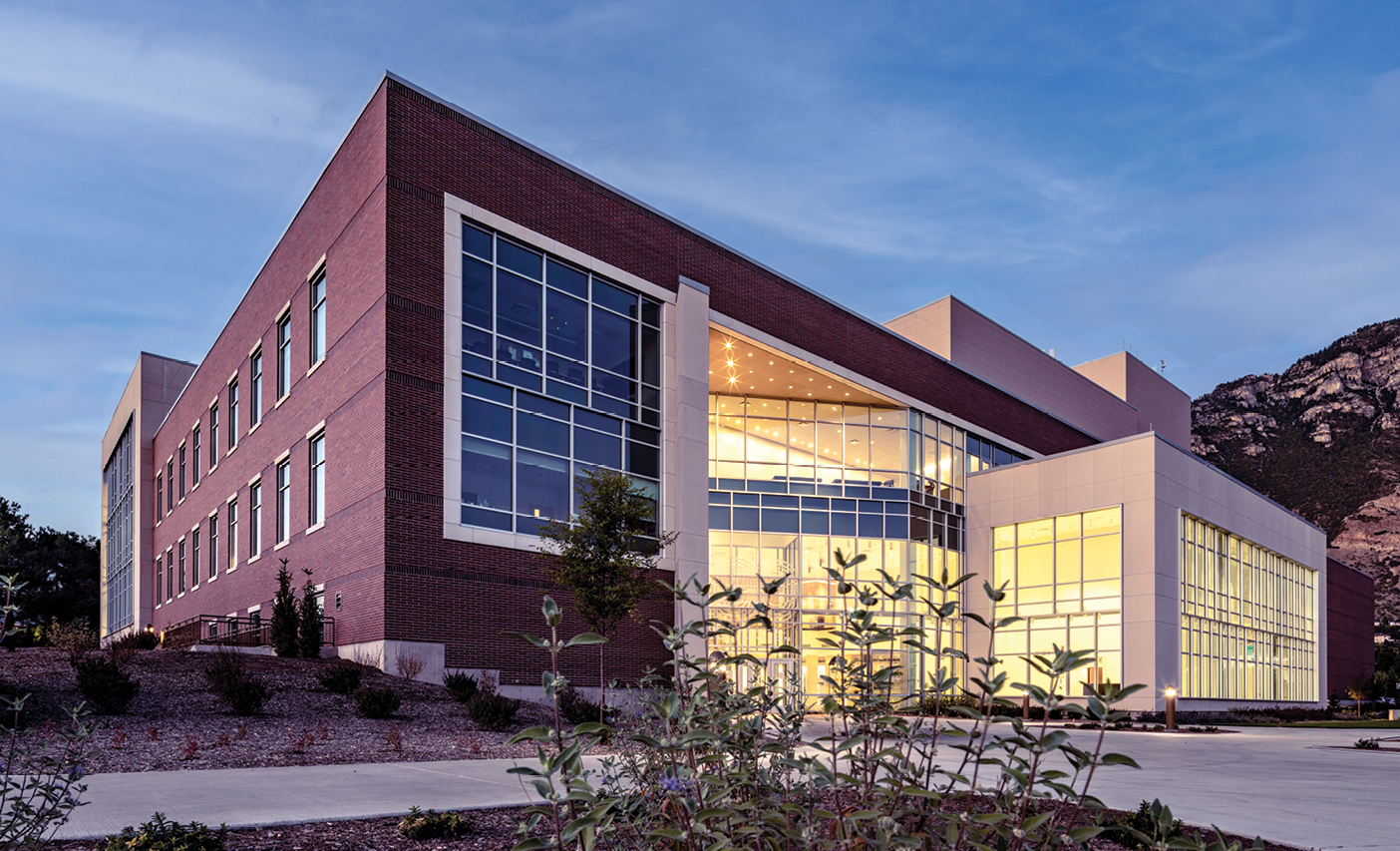 The new BYU Arts Building combines red brick, large glass windows, and illuminated interior spaces. Landscaping fills the foreground, and a rocky mountain rises behind the building beneath a blue evening sky.