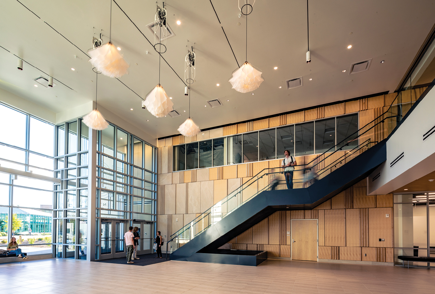 A spacious, sunlit atrium features floor-to-ceiling windows, light wood paneling, and a wide dark staircase leading to an upper level. Several students and visitors stand near the entrance and stairs, while sculptural pendant lights hang from the high ceiling.