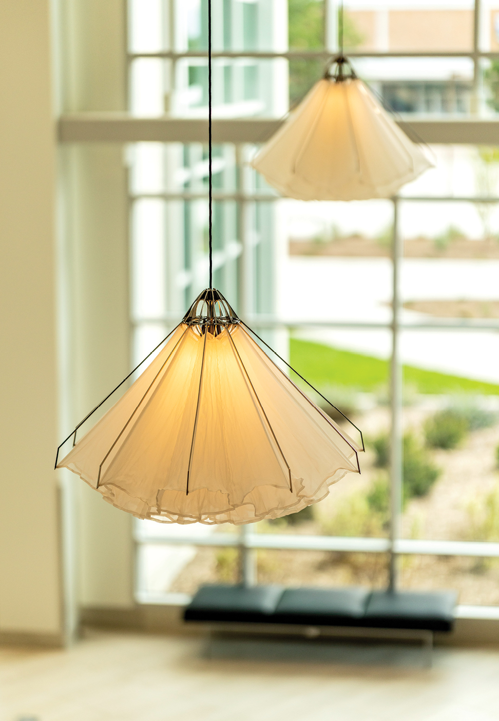 Two sculptural pendant lights hang in front of large windows in the Arts Building. Their translucent, pleated fabric shades resemble open flowers or jellyfish and glow softly in warm gold tones.