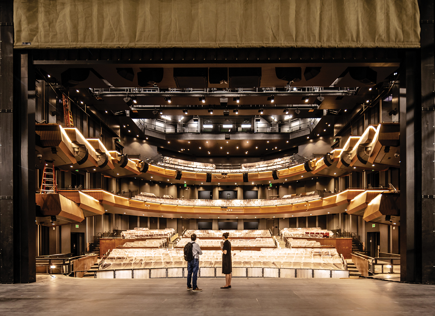 Two people stand on the stage of the unfinished Mainstage Theatre, facing rows of covered seats. The theater has multiple curved balconies, warm wood-colored railings, dramatic stage lighting, and an orchestra pit in front of the stage.