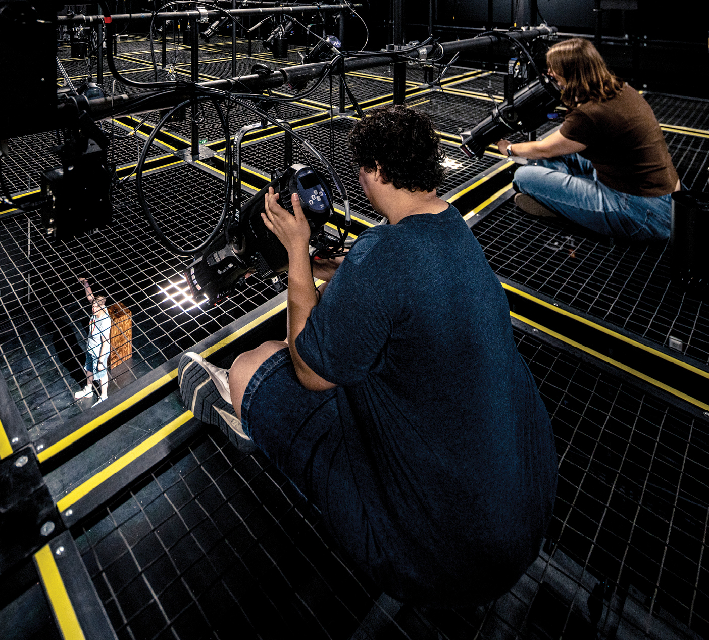 Two people crouch on a metal wire grid above the Black Box Theatre, surrounded by stage equipment, cables, and lighting fixtures. Yellow safety markings outline the walkable grid sections as they work with theater technology overhead.
