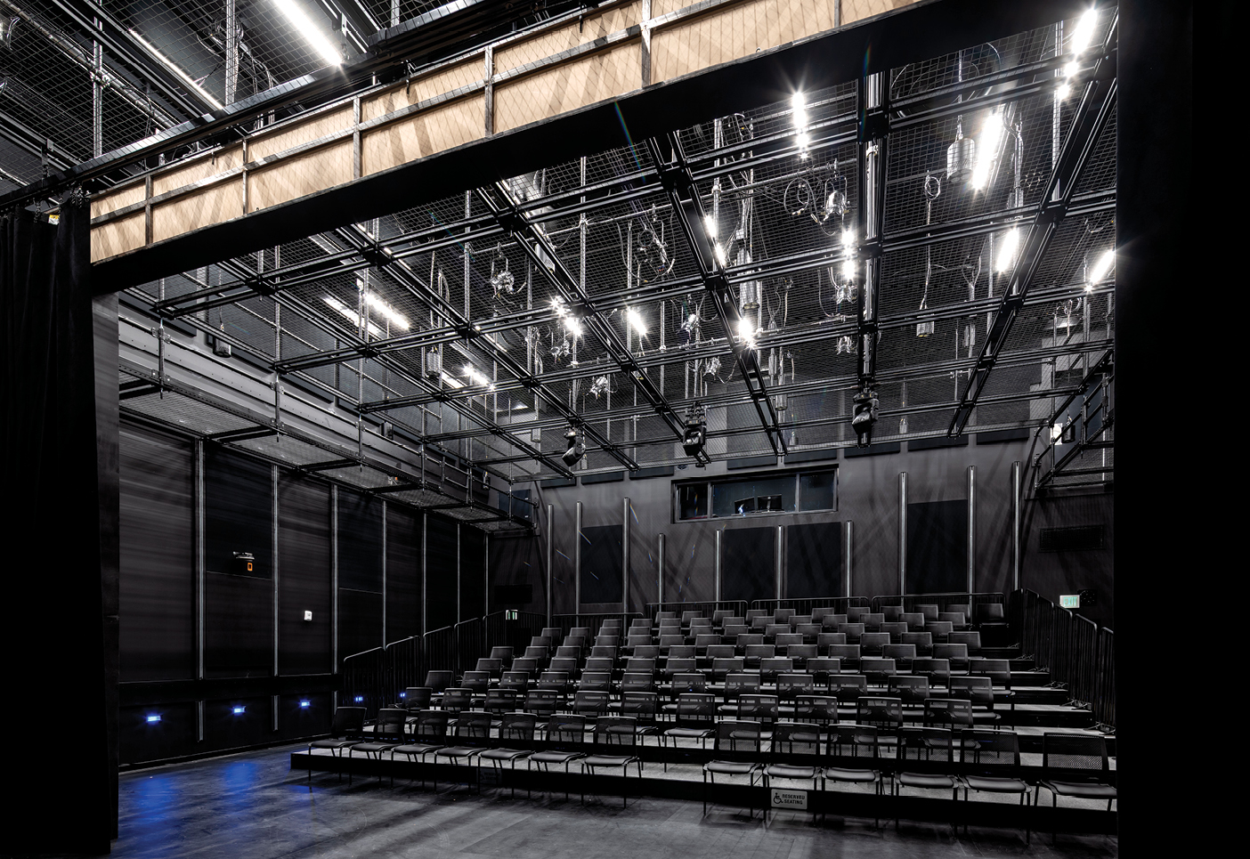 An empty, flexible 99-seat Black Box Theatre has rows of gray chairs arranged on a dark floor. Black walls and an overhead lighting grid create a simple, adaptable performance space, with small blue floor lights glowing along the sides.