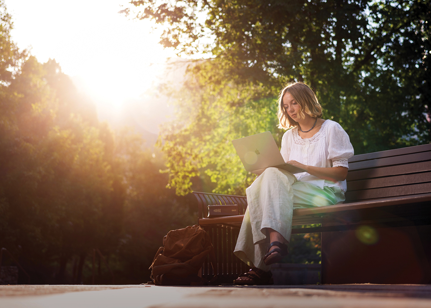 A woman sits on a bench and looks at her computer, illuminated by a ray of sunlight.