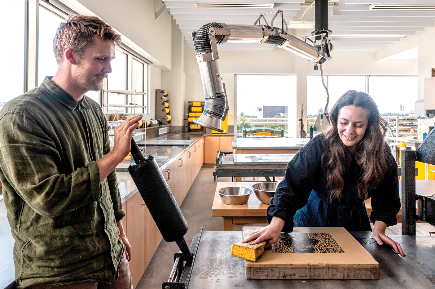 In the BYU Arts Building, Jennifer A. Watson (BFA ’06) and J. Eli Kerns (MFA ’26) prepare to print in a well-equipped studio, including Seussian ducts that safely whoosh away fumes from the room.