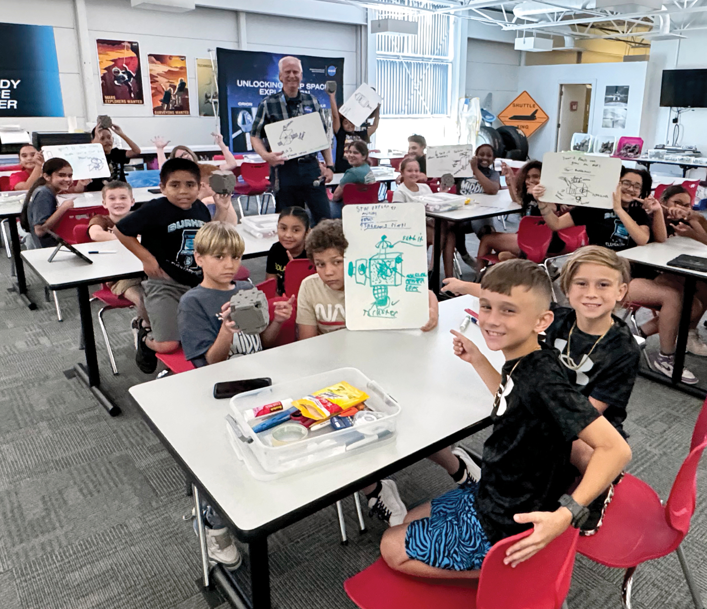 Students sitting at desks in classrooms holding up their satellite designs.