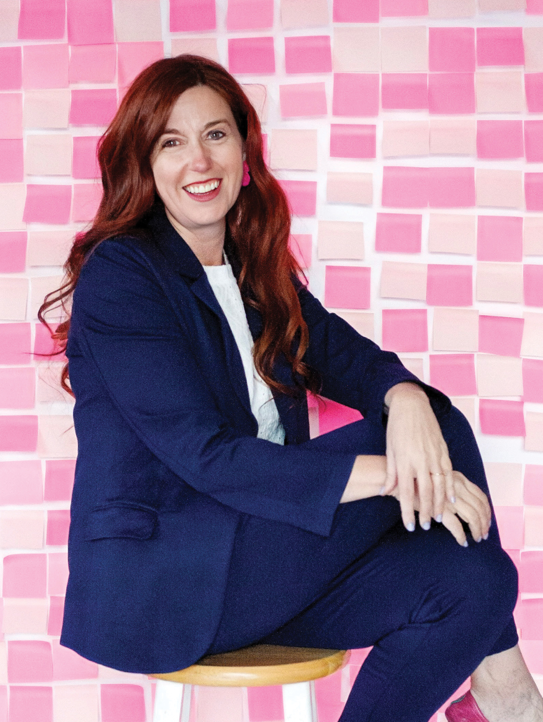 Kristen Hill Duke, a BYU alumna who coaches parents on parenting teens, smiles while seated on a wooden stool in front of a pink-and-white checkerboard wall. She has long auburn hair and wears a navy suit over a white shirt, with one arm resting on her raised knee.