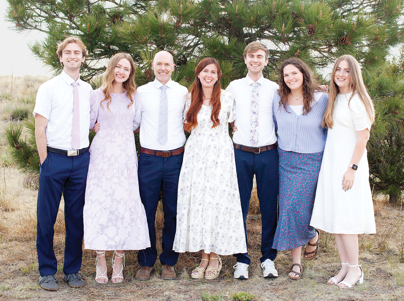 Kristen Hill Duke stands outdoors with her husband, Michael S. Duke, and several family members. The group poses beside a pine tree in a grassy setting, with the adults and young people dressed in light-colored, semi-formal clothing. The family photograph reflects Duke’s personal experience with parenting teens.