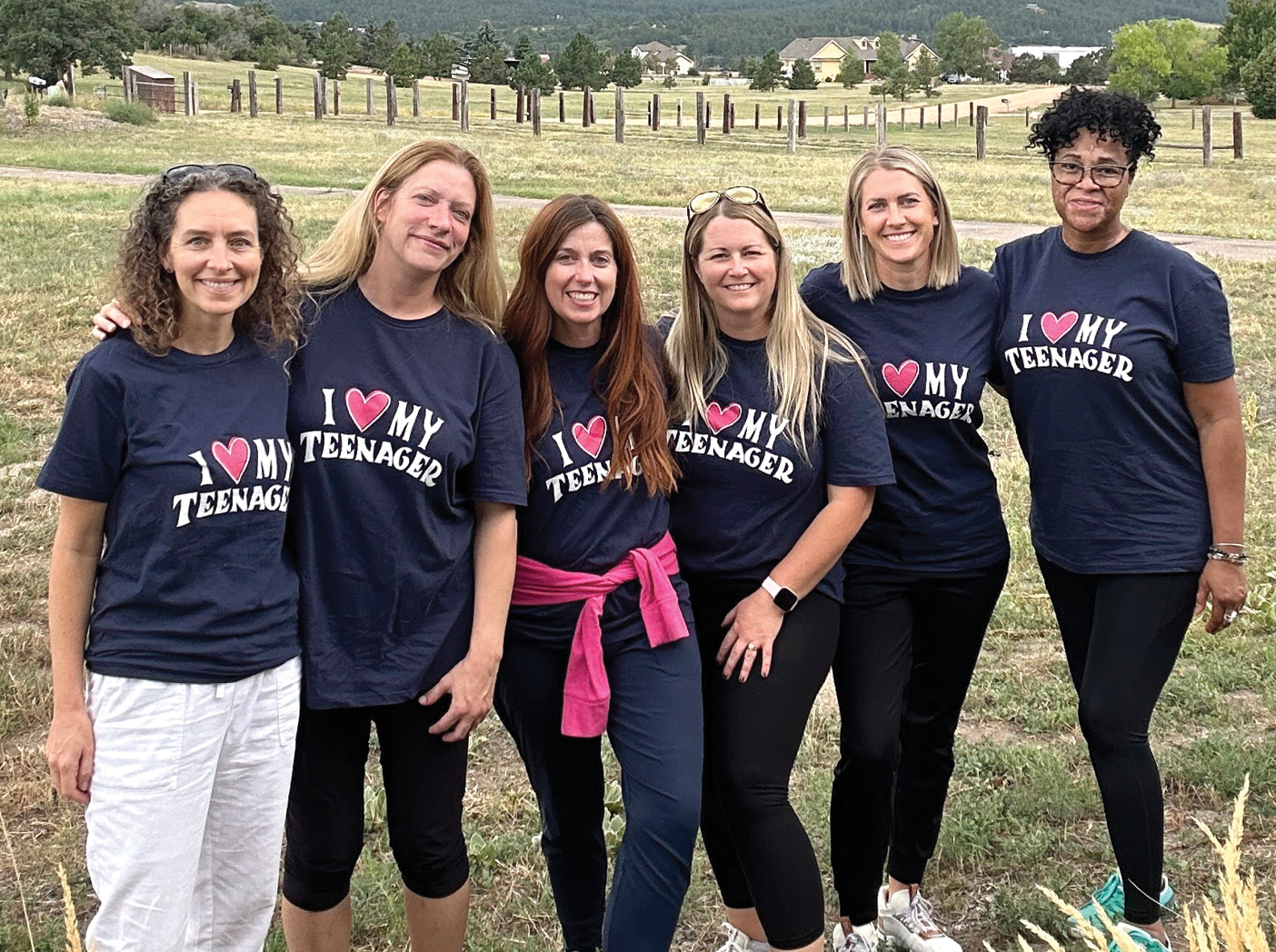 Six mothers of teenagers stand together outdoors at a TRUSTED retreat. They wear matching navy shirts with pink-and-white lettering that reads “I ♥ MY TEENAGER,” while posing in a grassy field with trees and a fence in the background.