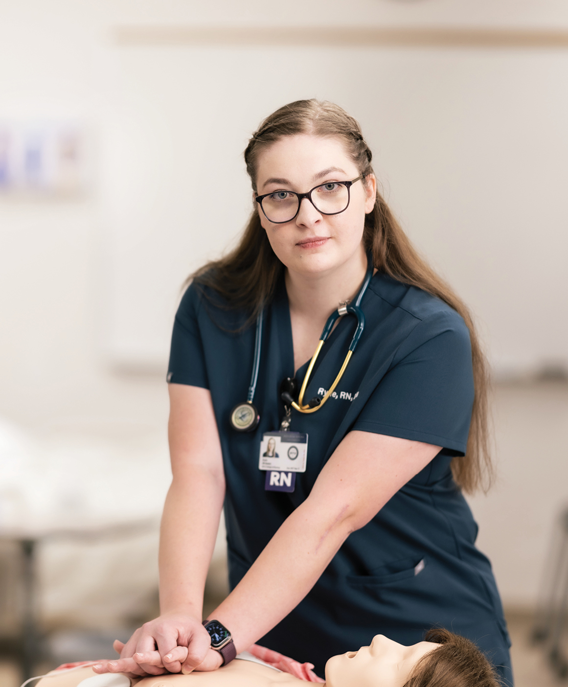 Rylie Rasmussen is in her scrubs facing the camera as she stands over a CPR manikin. In her pose, she ready to start performing CPR. 
